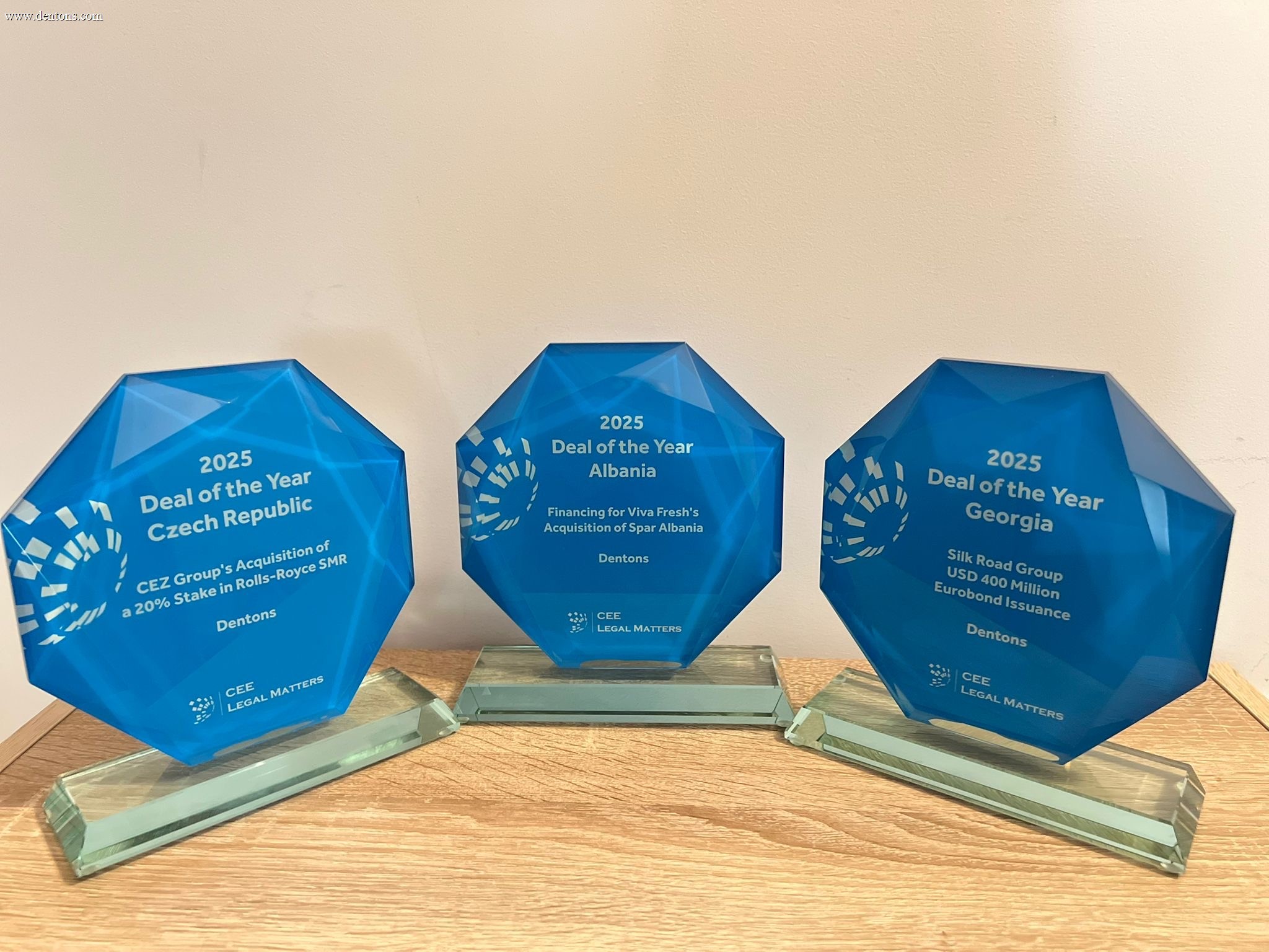 Three “Deal of the Year 2025” awards for Czech Republic, Albania, and Georgia, presented to Dentons by CEE Legal Matters, standing side by side on a wooden surface.